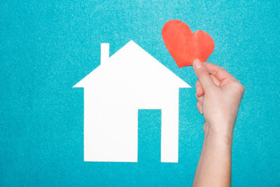 concept of love in family and at home. hand holds red heart over white paper house on blue background. Insurance of real estate. Loving and protecting the house.